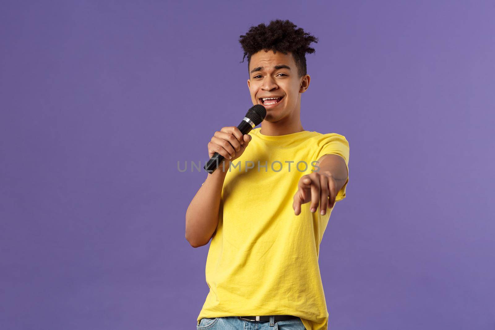 This song is for you. Portrait of romantic carefree hispanic man singing karaoke, pointing at camera as dedicate his performance, holding microphone, standing purple background.