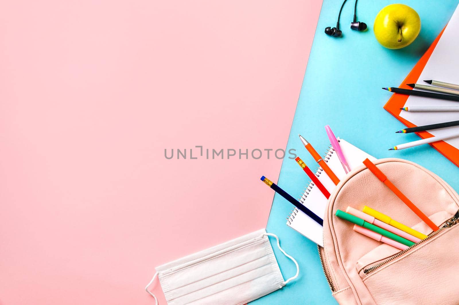 Creative student desk with protective mask, colorful modern school supplies and pink backpack on blue and pink duotone background. Top view. Flat lay. Copy space. Back to school concept