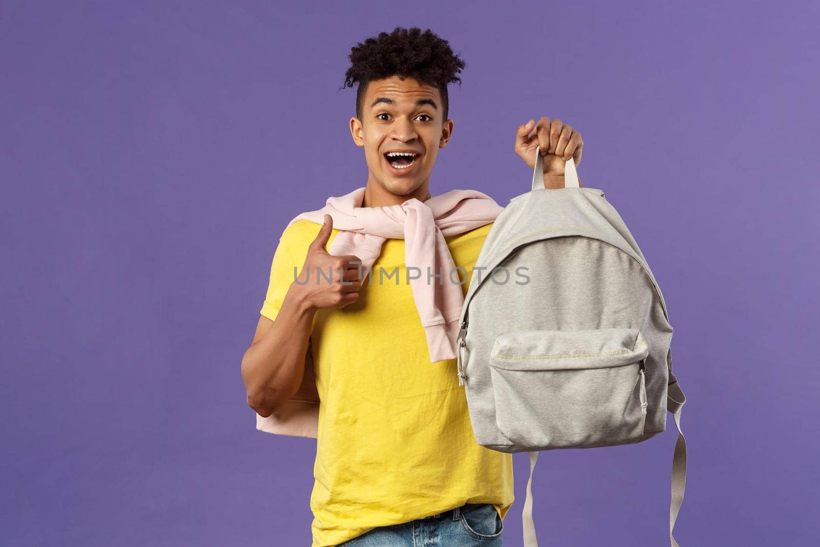 Portrait of cheerful young handsome male student, man recommending backpack, holding rucksack and show thumbs-up, bought new equipment for university semester, purple background.