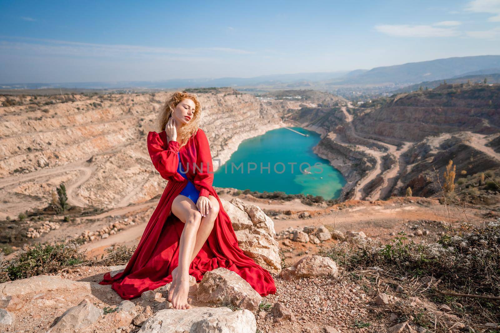 A beautiful girl in a red long dress, Sits on a rock high above the lake in the afternoon. Against the background of the blue sky and the lake in the shape of a heart. by Matiunina