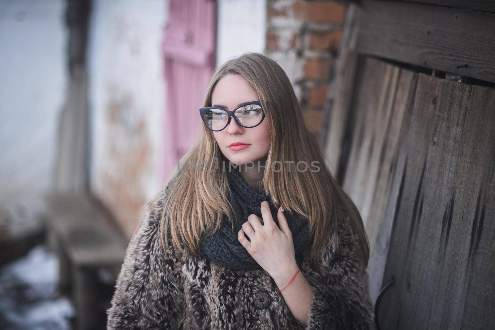 Girl blonde in cat eyes glasses in an artificial faux fur coat posing. Lying snow on the background. Red orange pink tones in the photo. Around the old shabby abandoned buildings. Cold winter