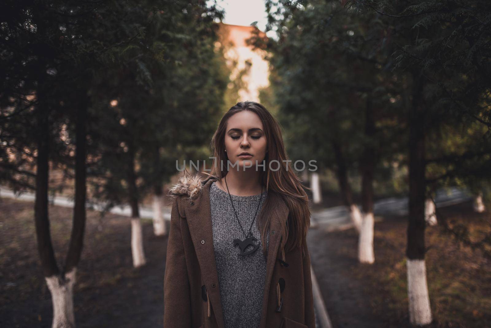 Woman in white pants and a brown coat near the trees by AndriiDrachuk