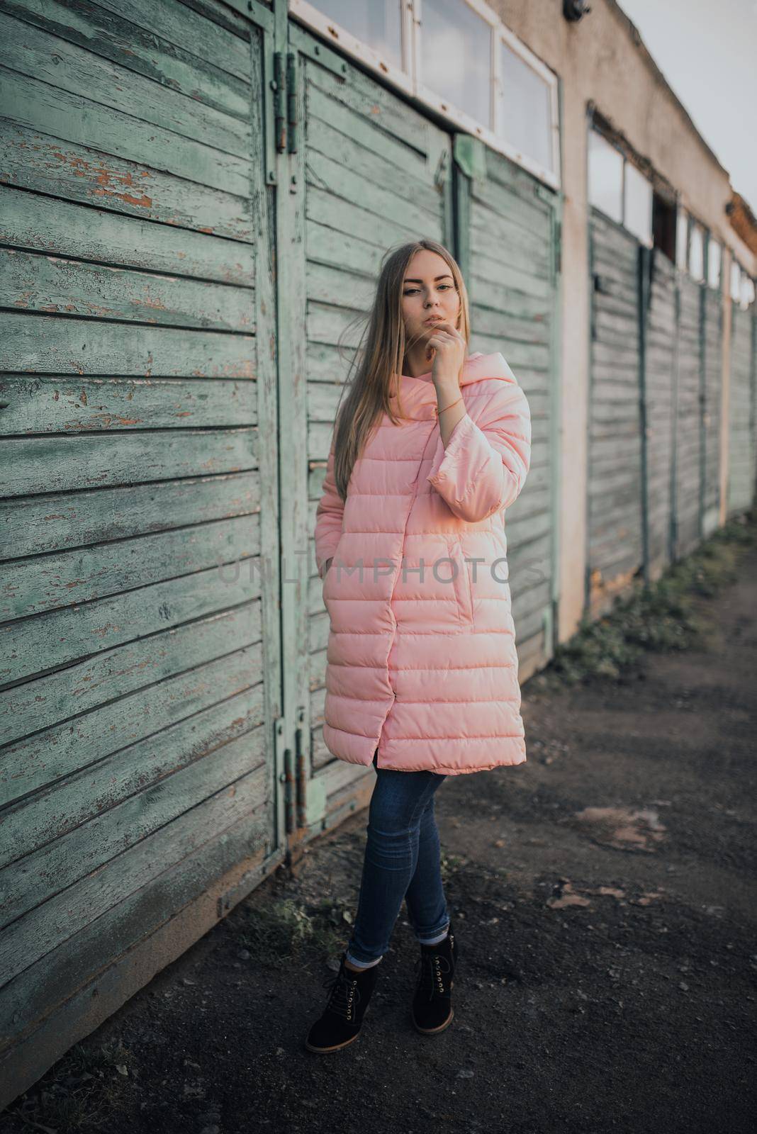 Young gorgeous blonde girl dressed fashion pink jacket and blue jeans. Old azure cerulean fence on the background. Picture ideal for illustrating women's magazines.