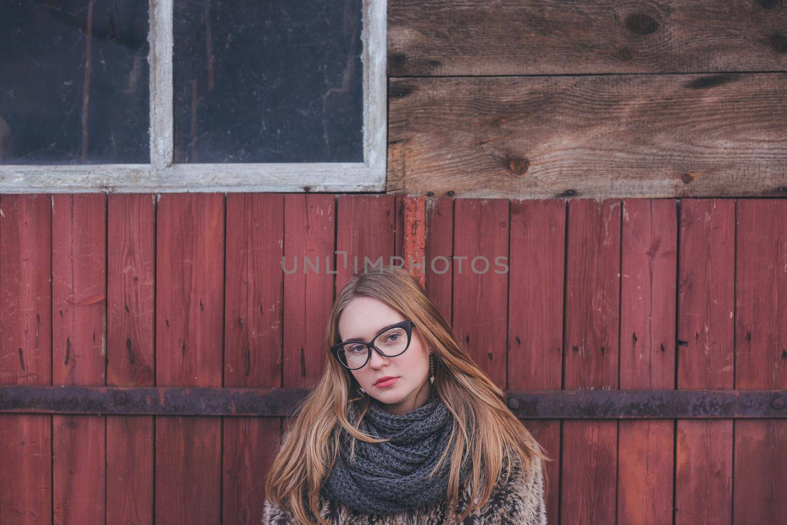 Girl blonde in cat eyes glasses in an artificial faux fur coat posing. Red orange pink tones in the photo. Around the old shabby abandoned buildings. Cold winter. wooden wall