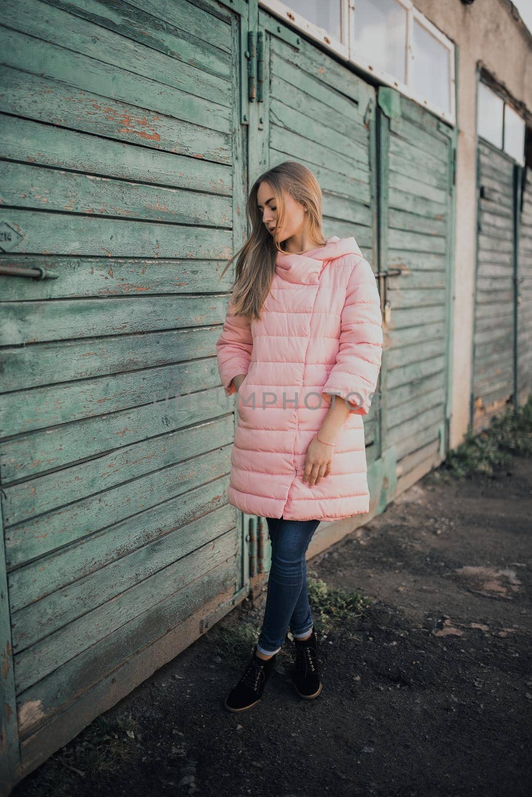 Young gorgeous blonde girl dressed fashion pink jacket and blue jeans. Old azure cerulean fence on the background. Picture ideal for illustrating women's magazines.