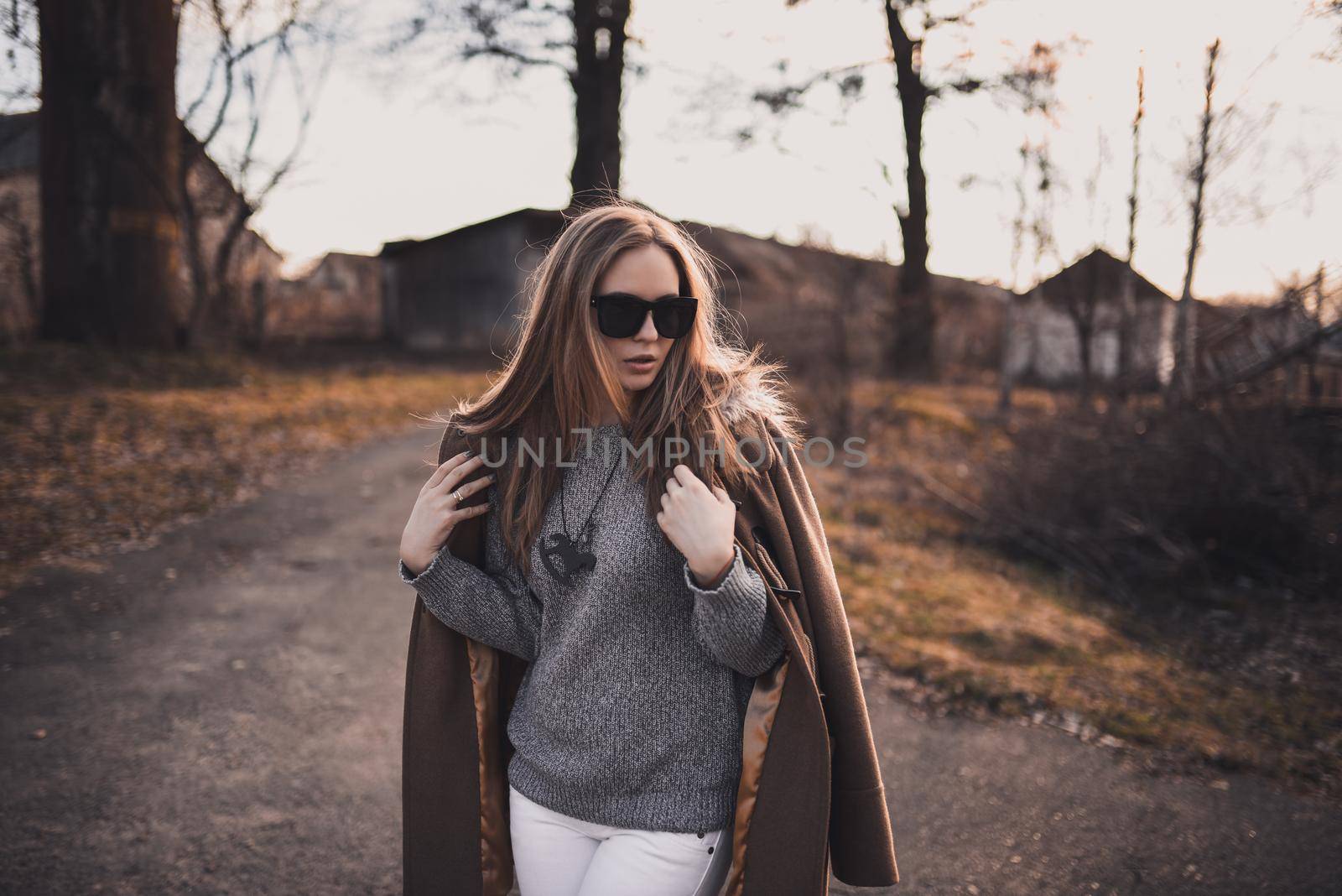 beautiful young blonde model girl. White pants. gray knitted sweater. black boots. black sunglasses. wooden pendant on the neck in the form of a horse. in a brown coat posing. On the Sunset. Portrait