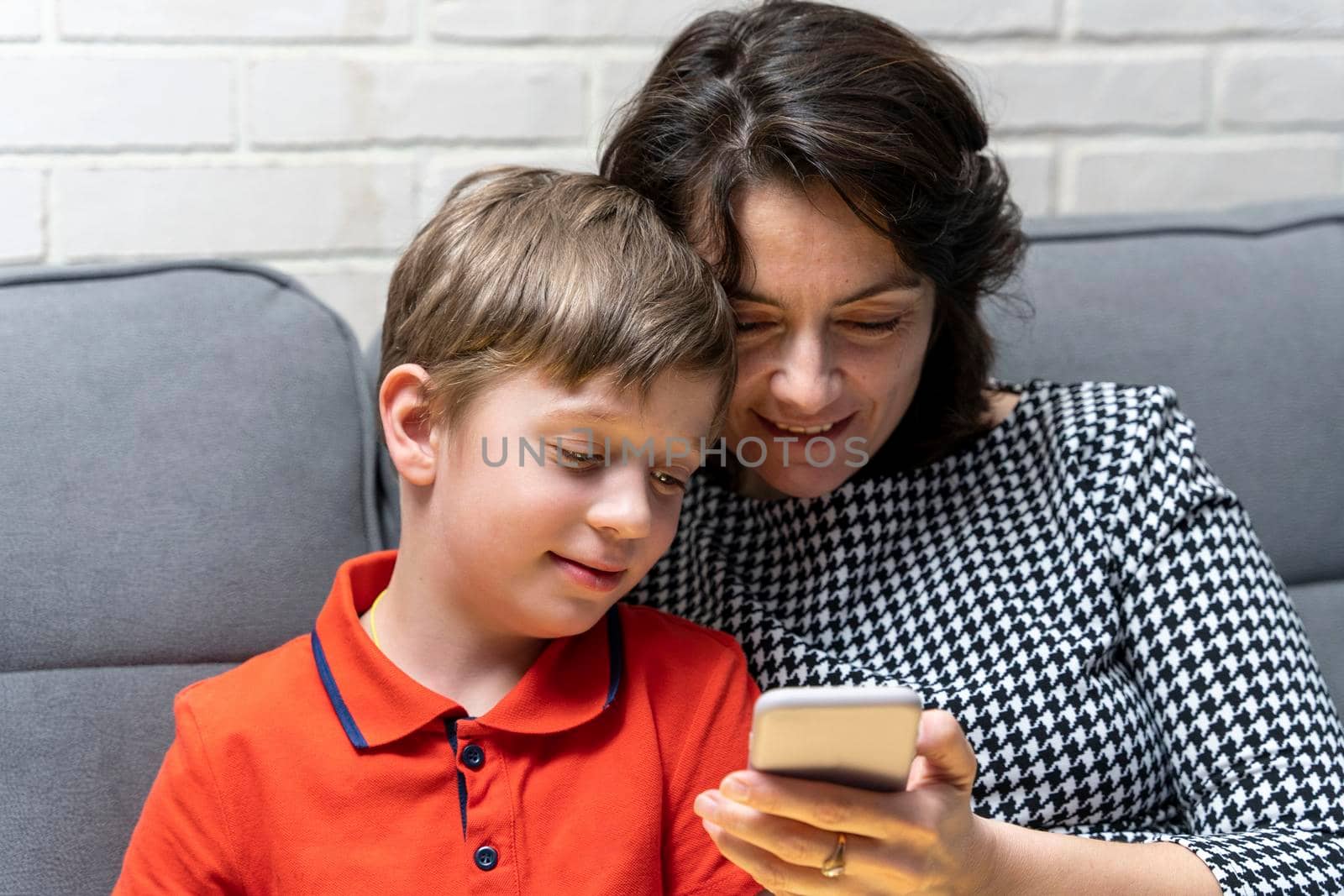 happy European preschooler boy and his mom are sitting on the couch and looking at the smartphone screen, reading congratulations, chatting online. Dependence on gadgets