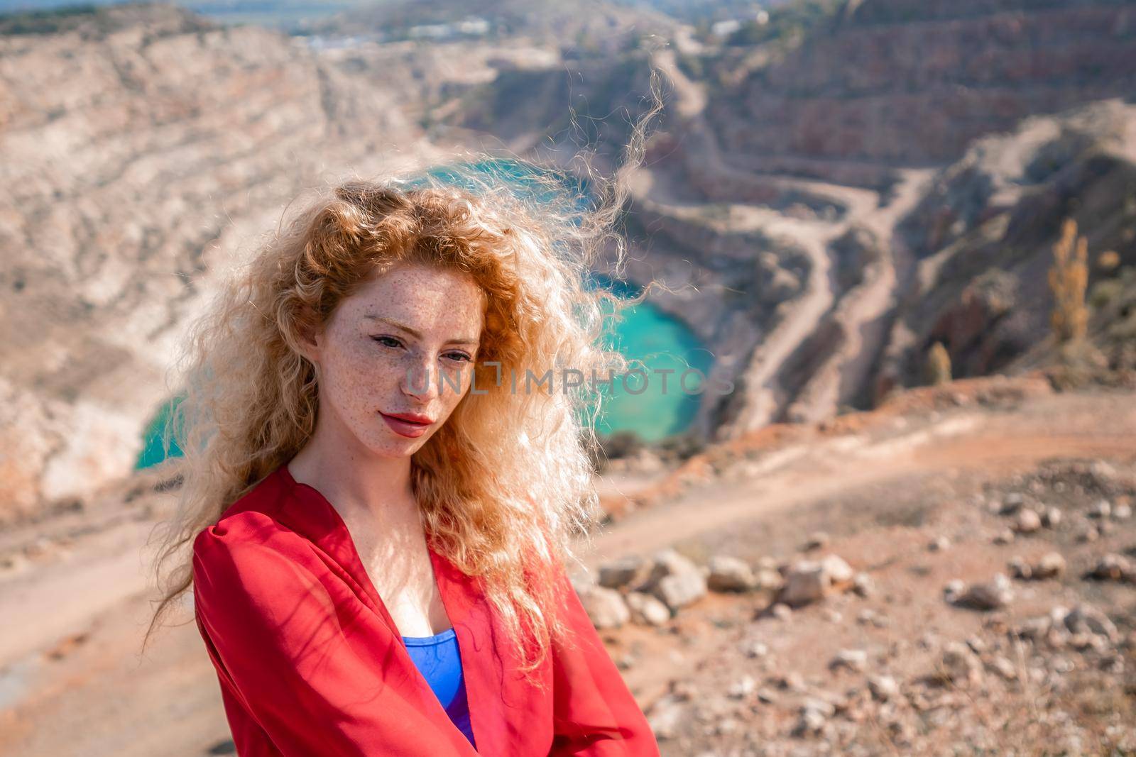 A beautiful girl in a red long dress, Sits on a rock high above the lake in the afternoon. Against the background of the blue sky and the lake in the shape of a heart