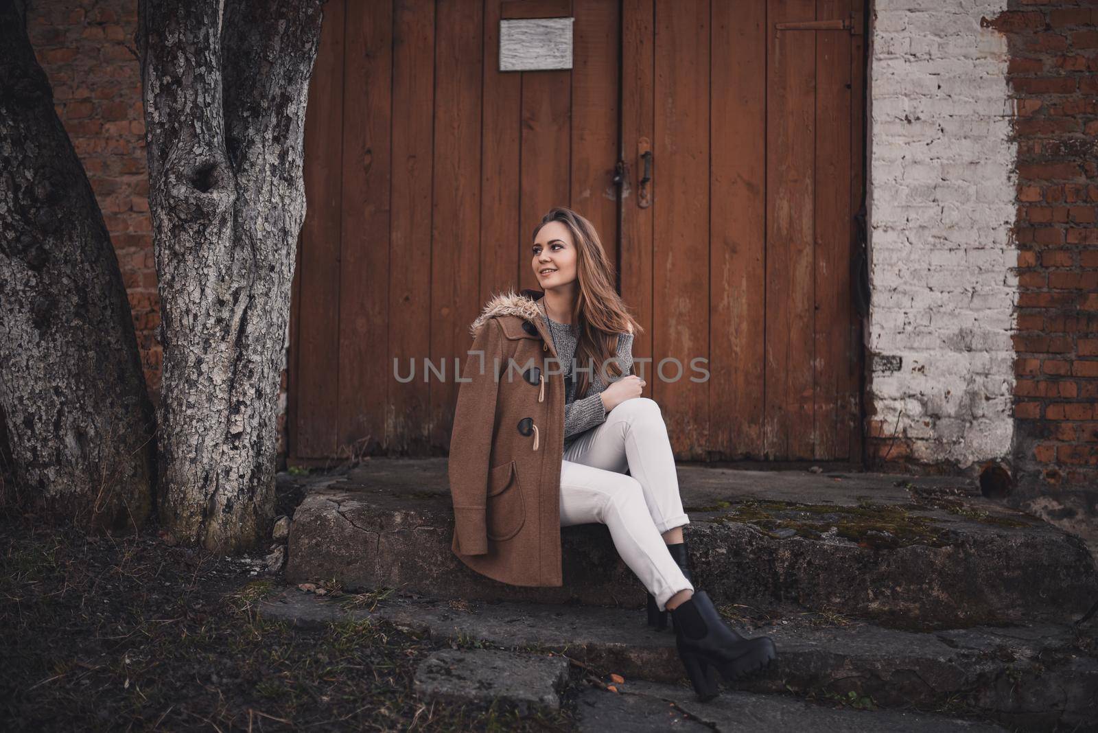 beautiful young blonde model girl. White pants. gray knitted sweater. boots. wooden pendant on the neck in the form of a horse. in a brown coat posing. On the Sunset. Portrait. sitting on the steps