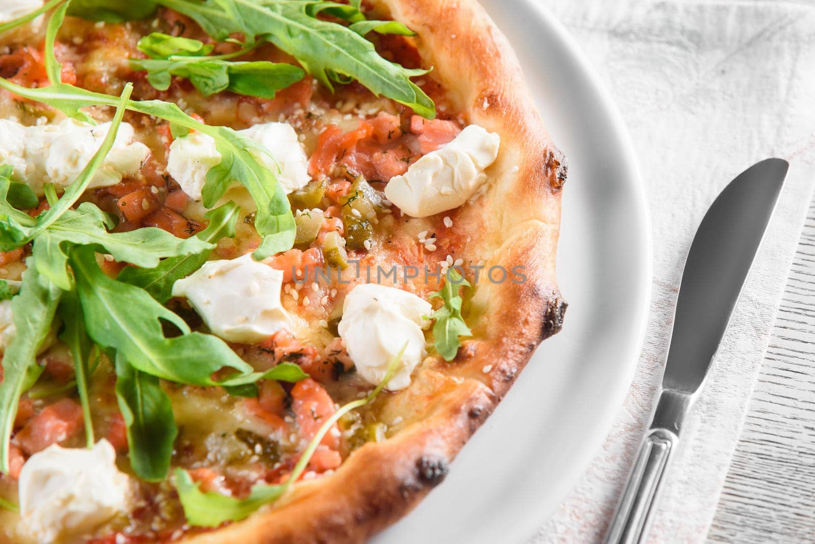 Pizza with salmon, feta cheese, sauce, tomatoes, on white round plate on light wooden table on background of glass of red wine.