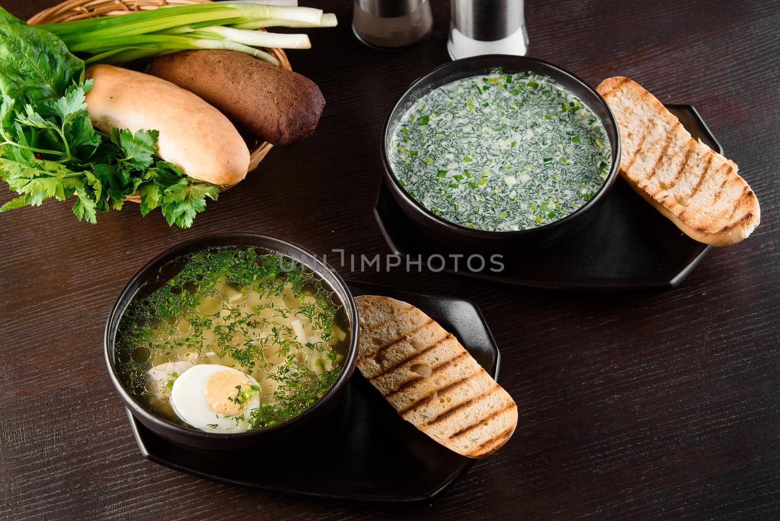 Okroshka, broth with noodles and eggs in black plate, toasted bread, herbs on dark wooden table. Healthy seasonal lunch