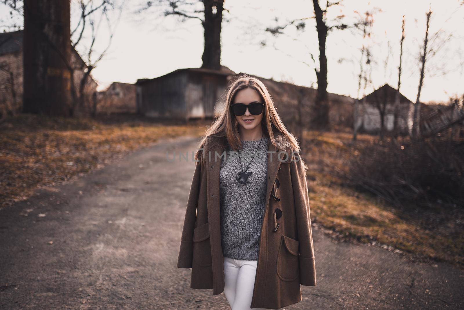 beautiful young blonde model girl. White pants. gray knitted sweater. black boots. black sunglasses. wooden pendant on the neck in the form of a horse. in a brown coat posing. On the Sunset. Portrait