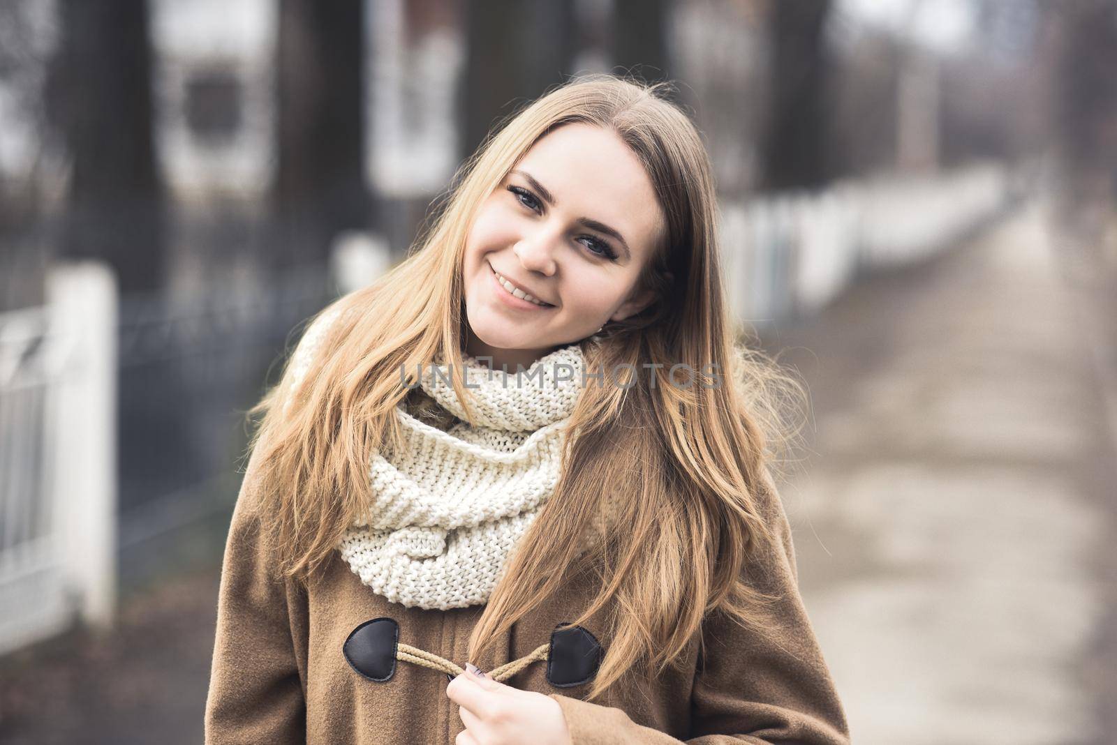 Blond-haired young girl cheerful joyful dressed you in a beige coat and white scarf. Autumn gray street on the background. empty road