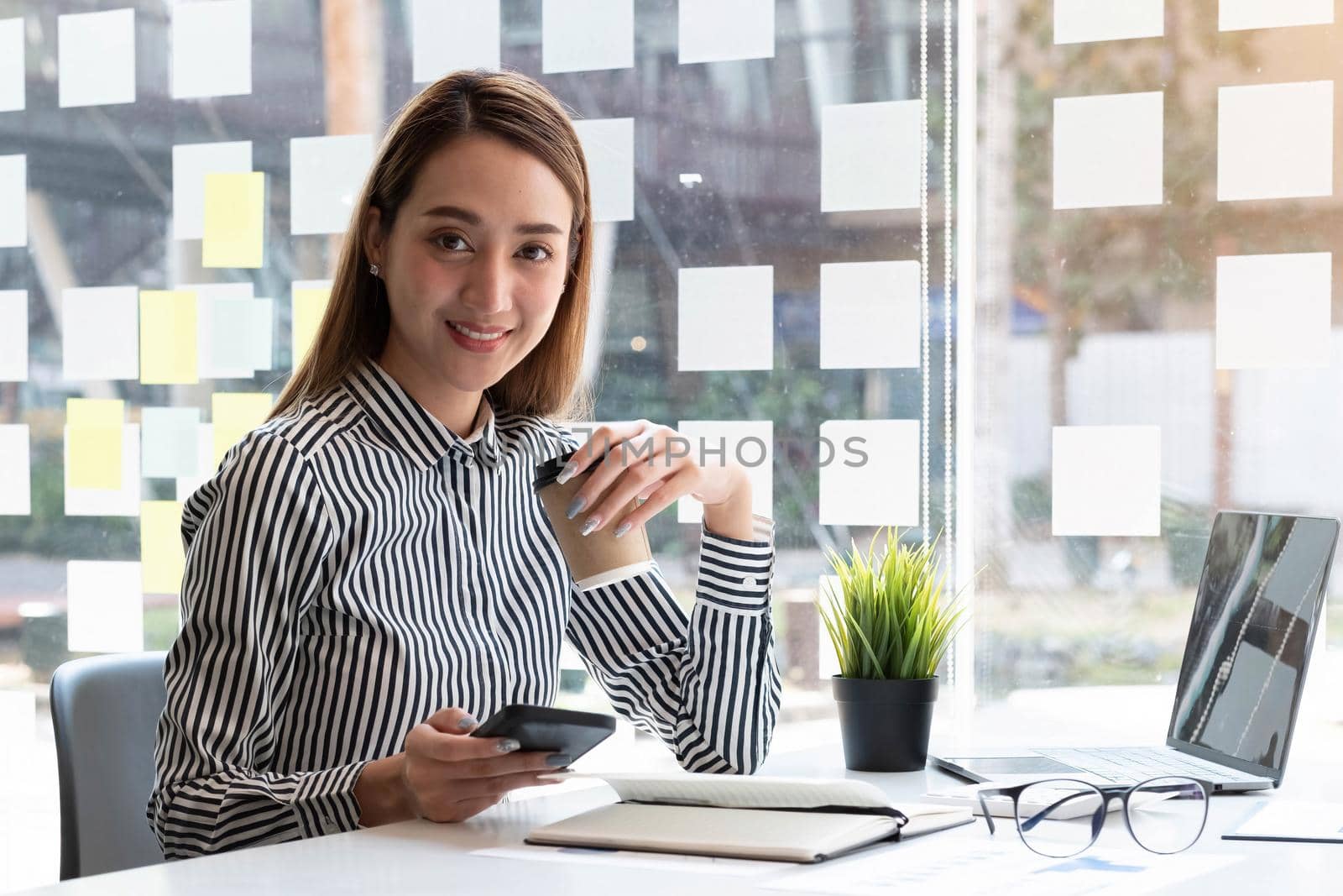 Beautiful young Asian businesswoman drinking a coffee and holding a smartphone working on laptop at office. Looking at camera. by wichayada