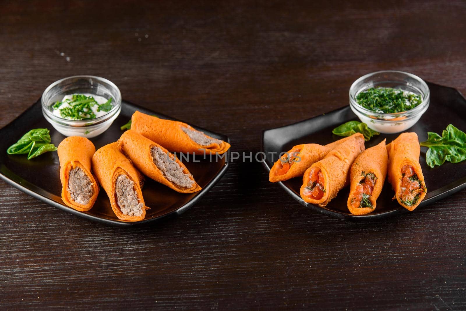 Orange pancakes with pate, salmon, dill, sour cream on black plate on a wooden table. Filled rolls.