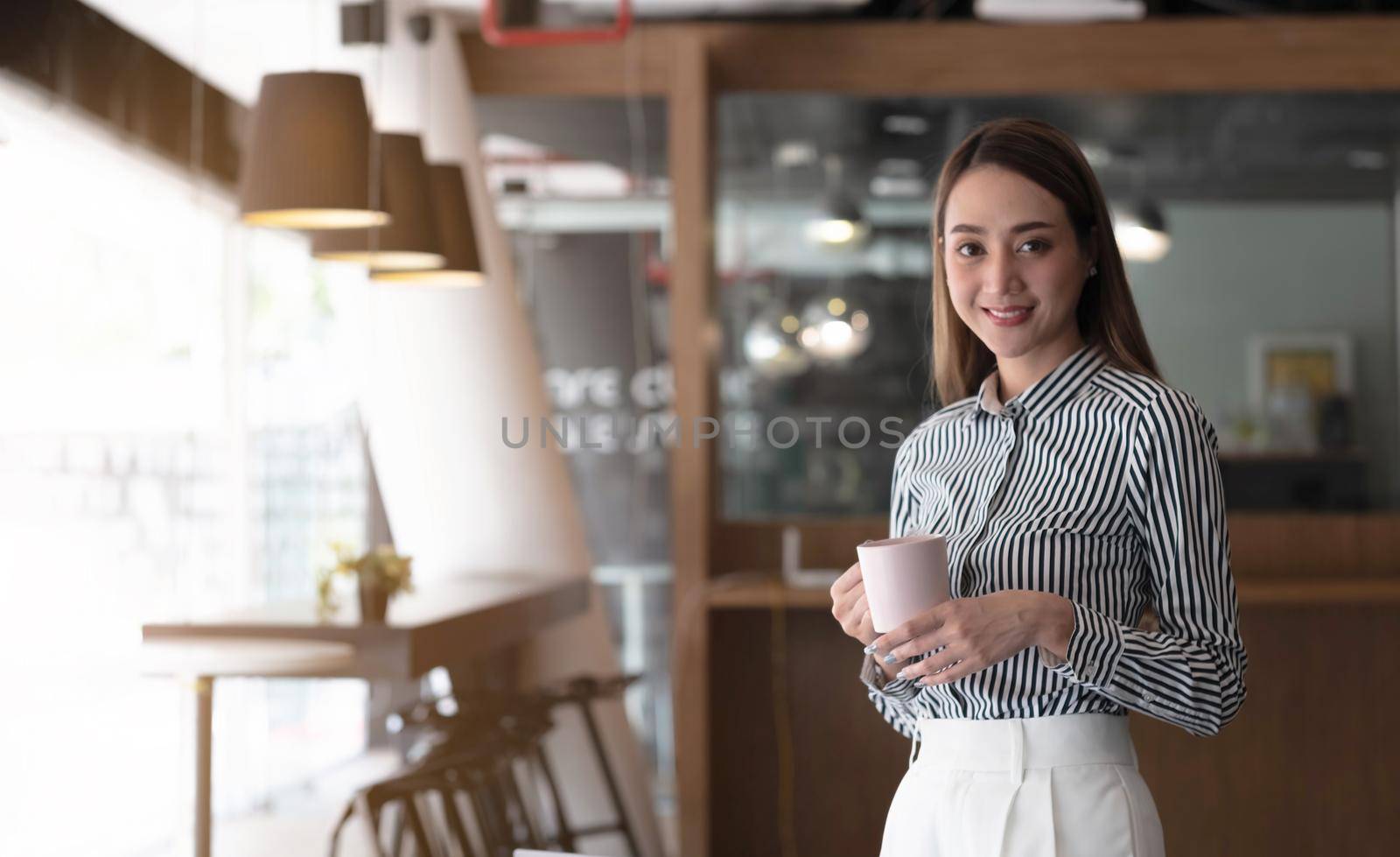 Smiling Asian businesswoman holding a coffee mug and laptop at the office. Looking at the camera. by wichayada