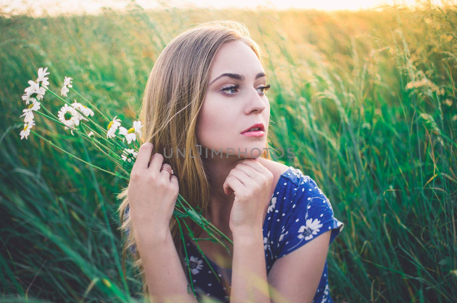 young slim fair-skinned girl blonde beautiful sit in tall grass. Girl in a blue short tight dress with a print of daisies. A bouquet daisies in hand.