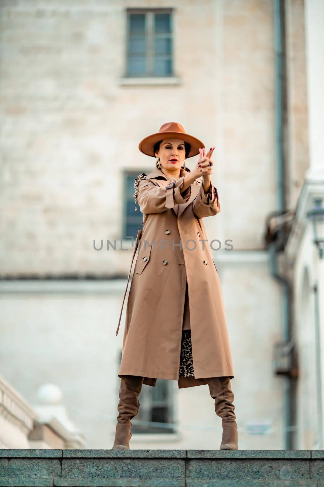 Outdoor fashion portrait of young elegant fashionable brunette woman, model in stylish hat, choker and light raincoat posing at sunset in European city. by Matiunina