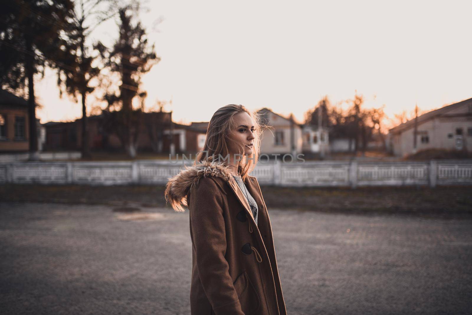 beautiful young blonde model girl. White pants. gray knitted sweater. black boots. . wooden pendant on the neck in the form of a horse. in a brown coat posing. On the Sunset. Portrait. near the trees