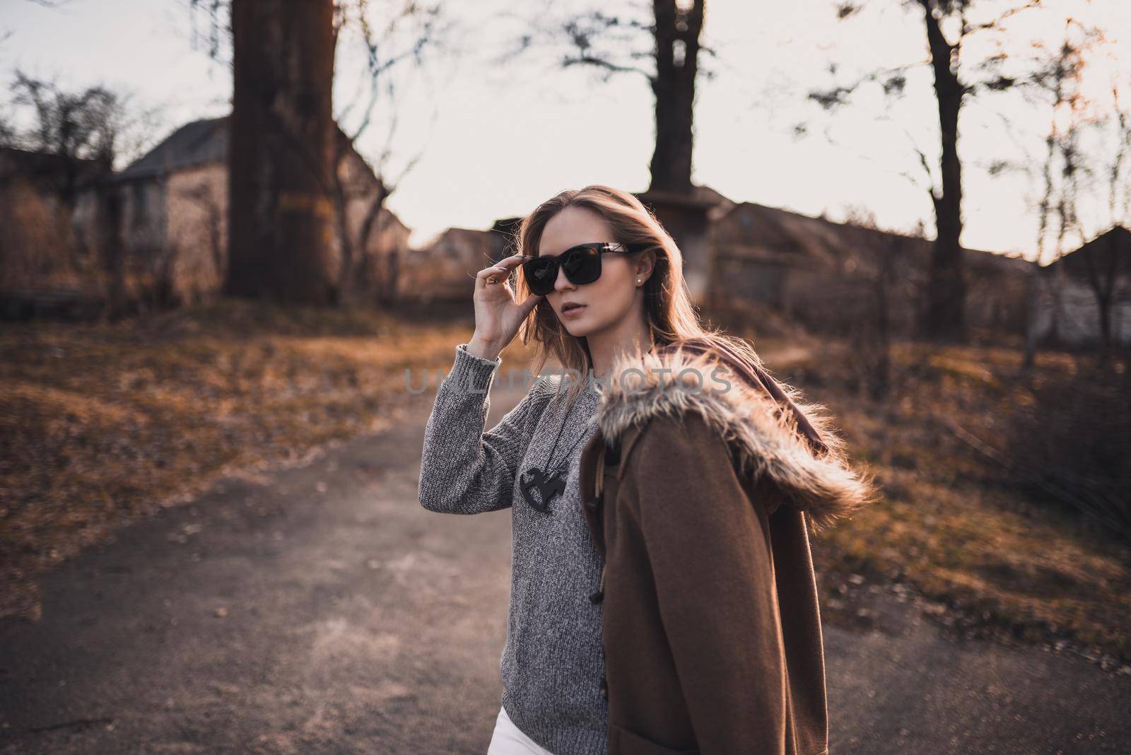 beautiful young blonde model girl. White pants. gray knitted sweater. black boots. black sunglasses. wooden pendant on the neck in the form of a horse. in a brown coat posing. On the Sunset. Portrait