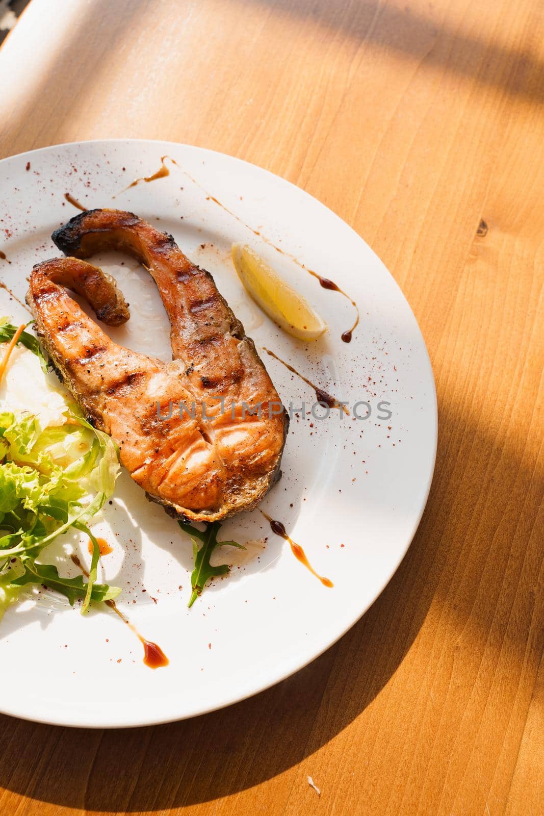 Grilled salmon steak with lettuce and lemon on white plate on wooden table by Rabizo