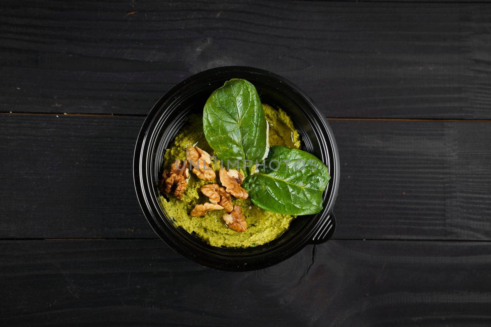 Hummus with basil and walnuts garnished with herbs in black bowl on dark wooden table.