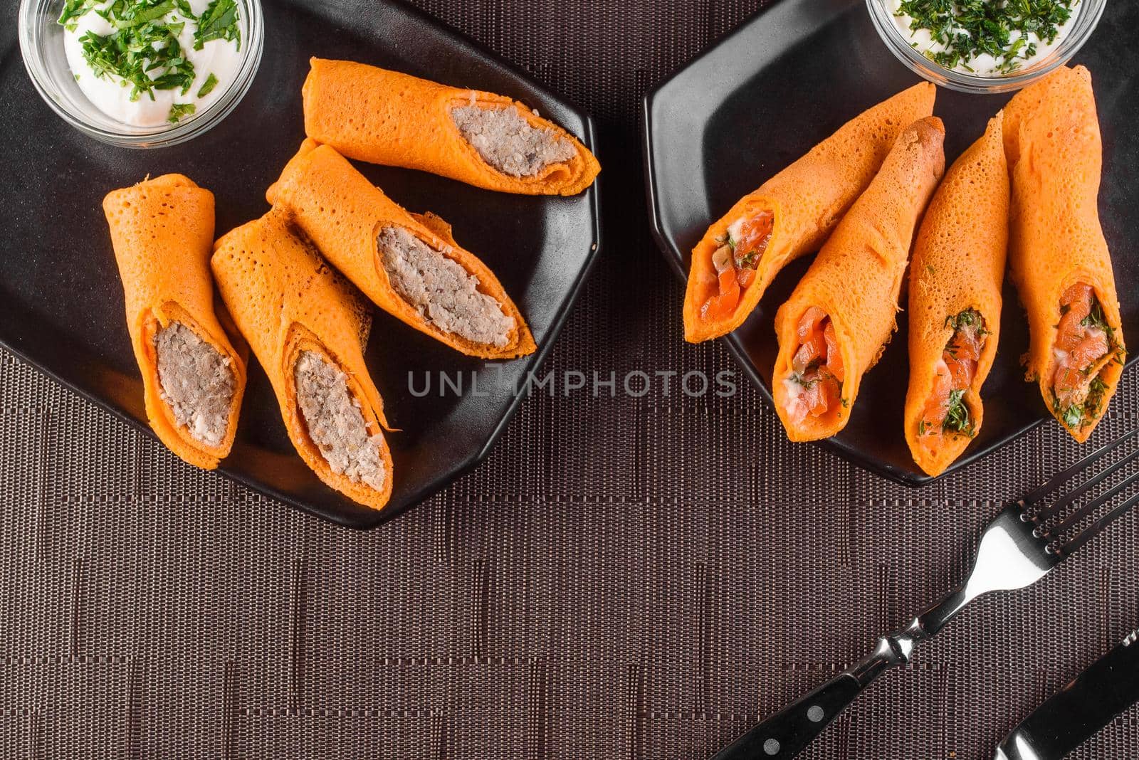 Orange pancakes with pate, salmon, dill, sour cream on black plate on a wooden table. Filled rolls.