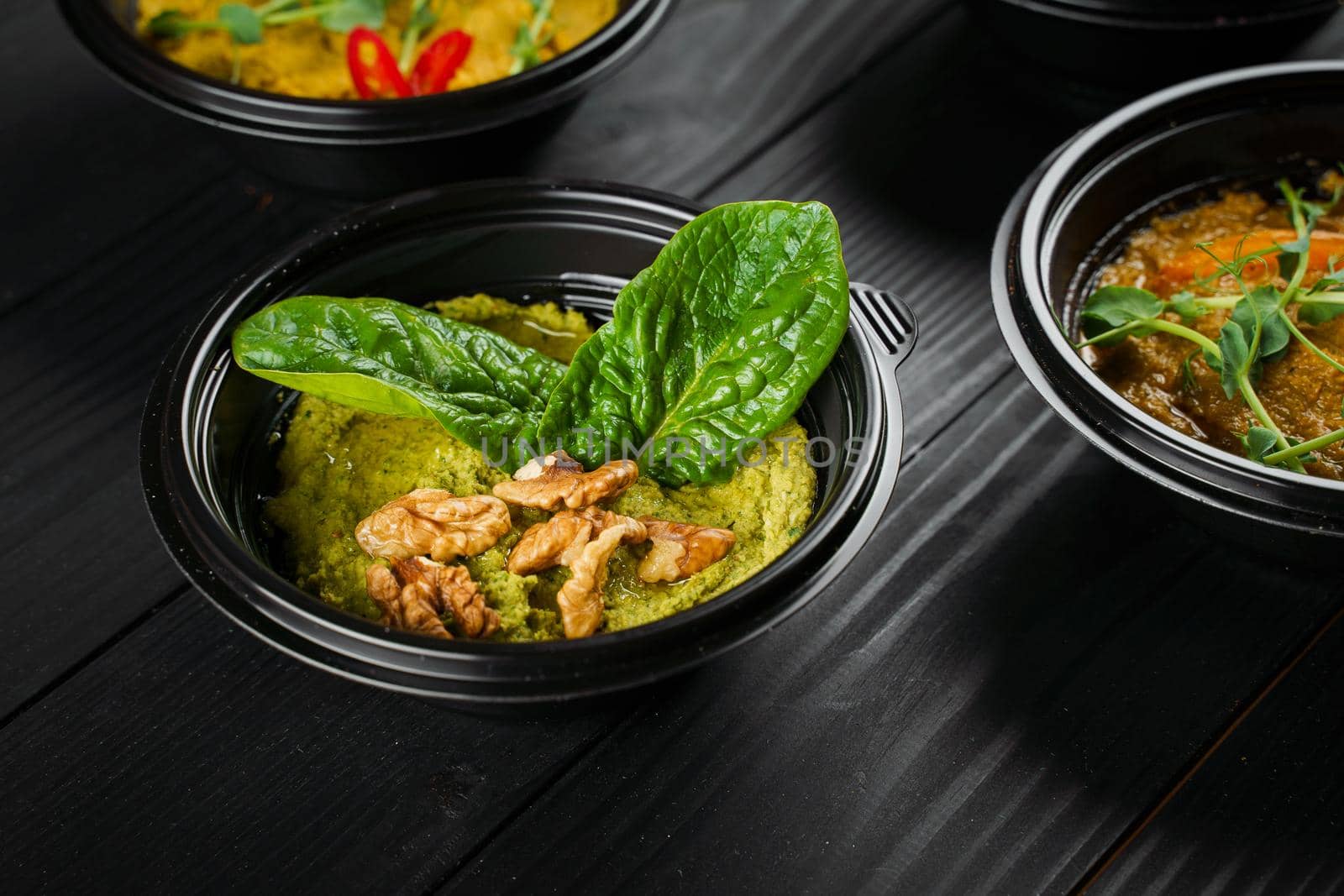 Hummus with basil and walnuts garnished with herbs in black bowl on dark wooden table.