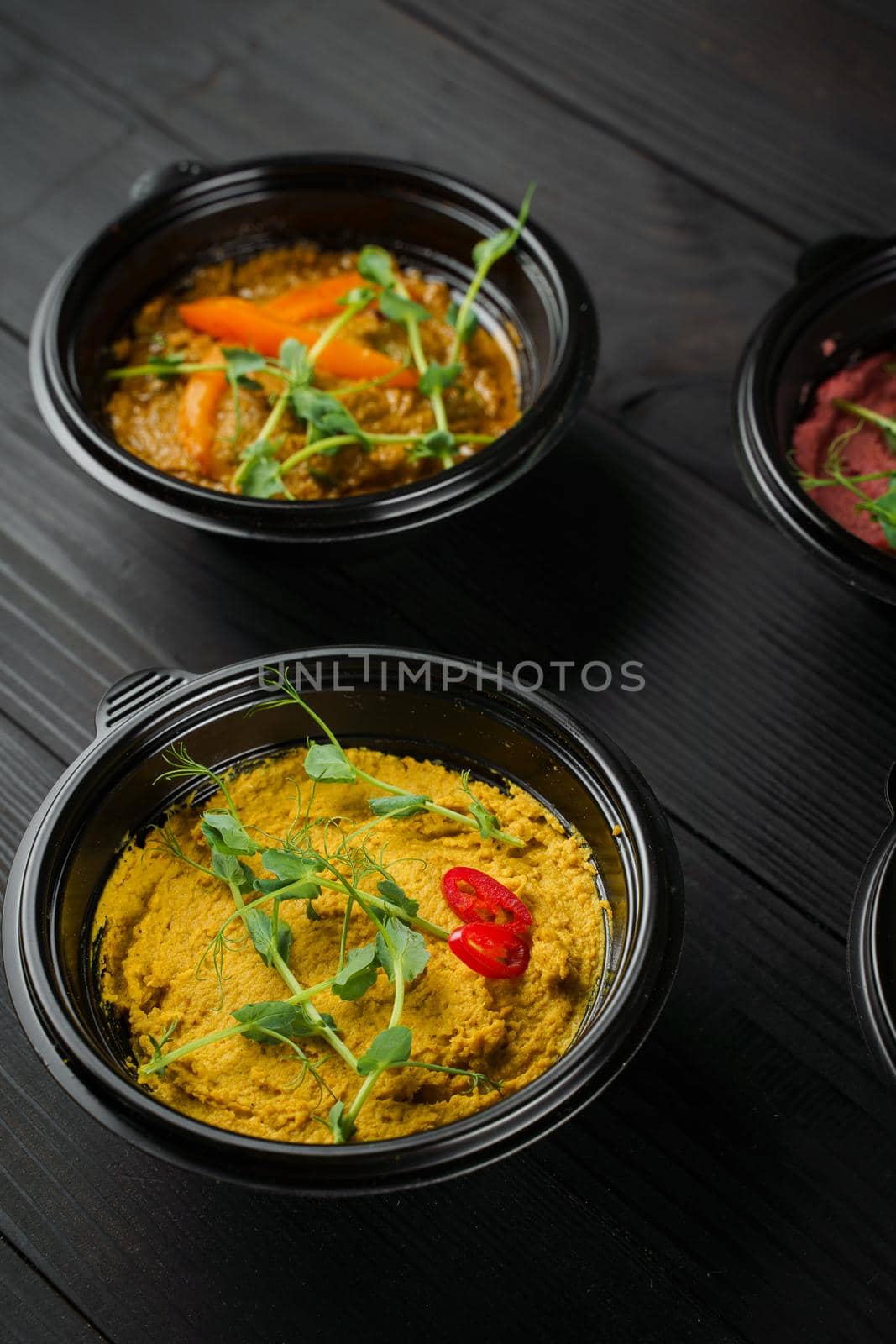 Hummus garnished peppers, chili, beet and herbs in black bowl on dark wooden table. Hummus assortment by Rabizo