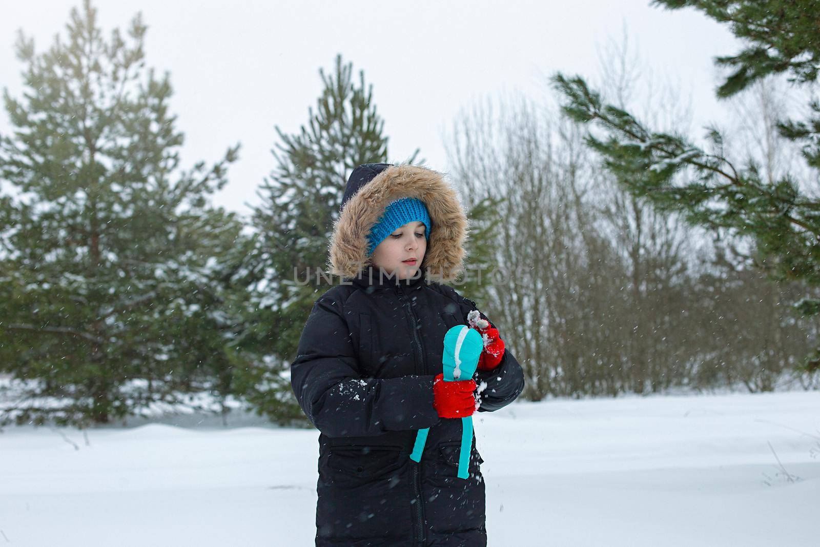 A teenage boy in a black jacket and a striped knitted hat, in winters park , makes snowballs using a light blue plastic sculpting tool. Close up. Copy space
