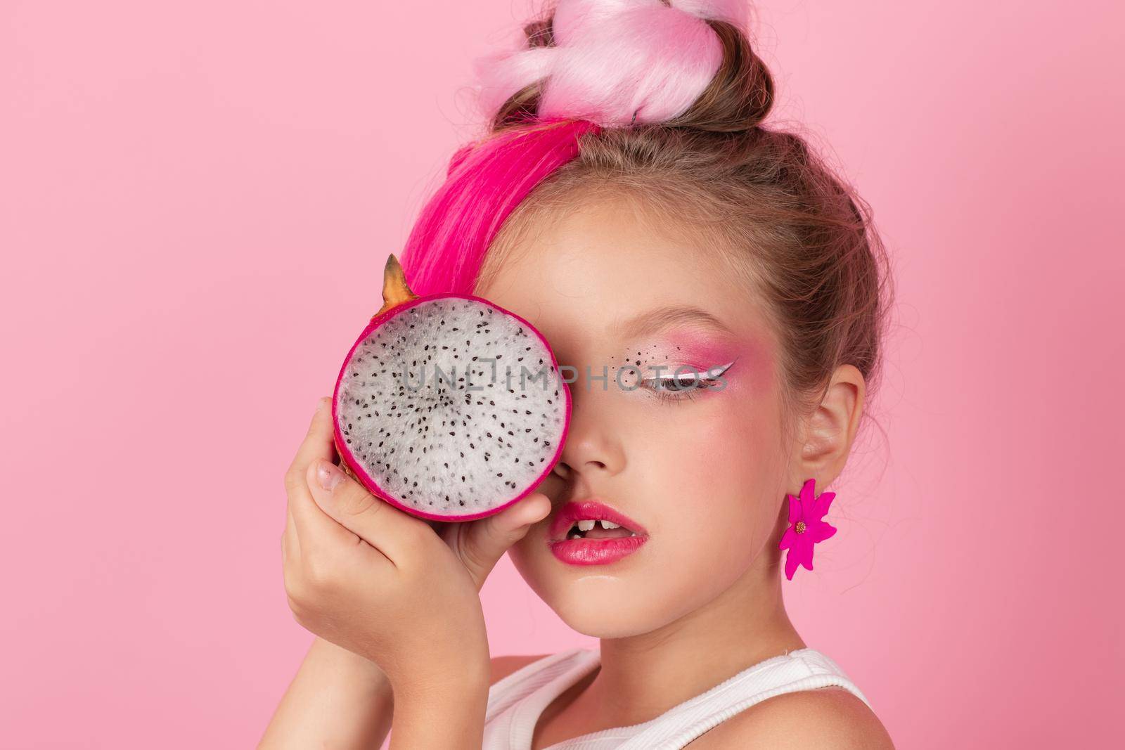 Close-up portrait of pretty girl with pink hairstyle with dragon fruit on pink background. Studio shot of charming tween girl with pink make up enjoying juicy red pitaya. exotic Pitahaya fruit.