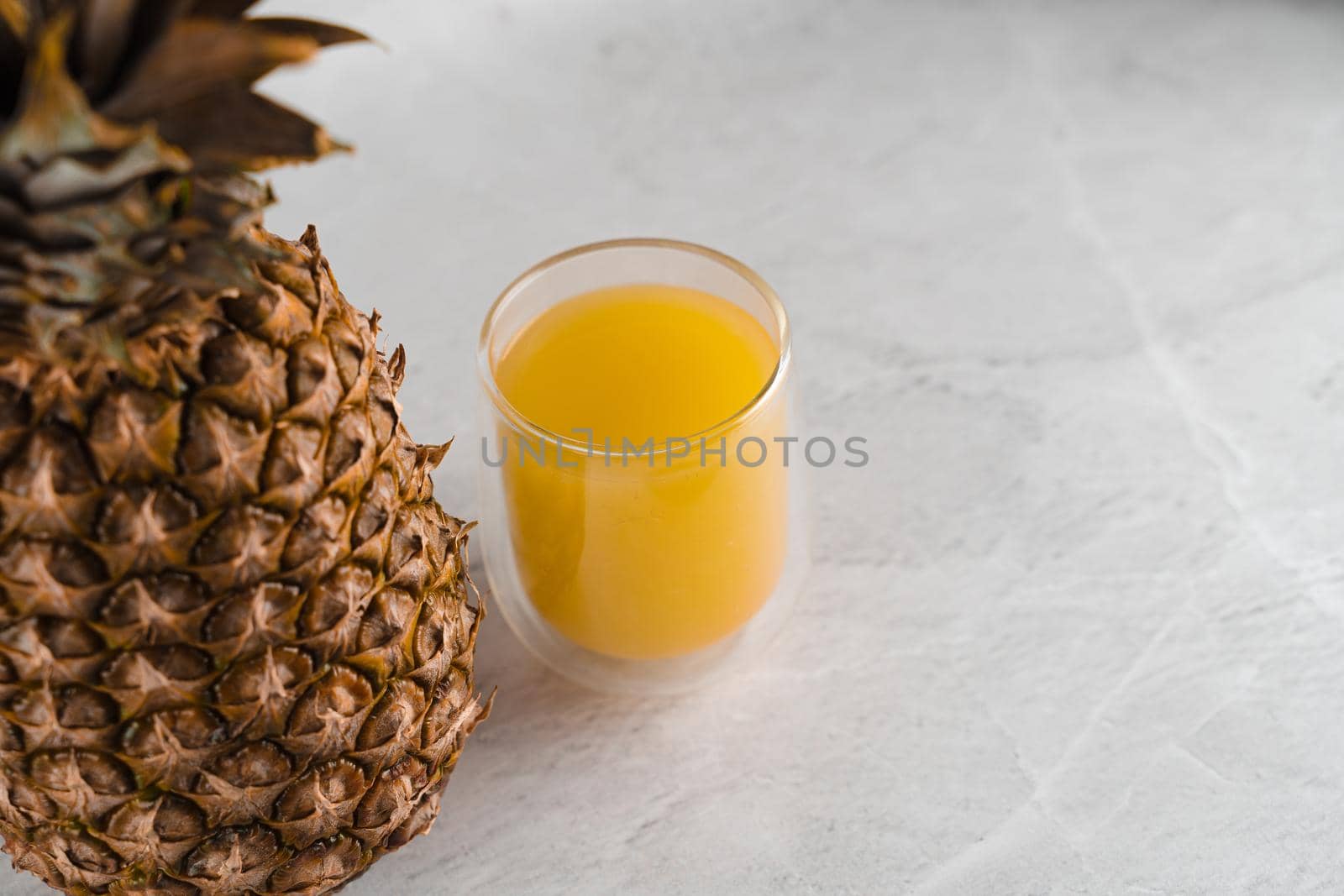 Pineapple fruit and juice in double glass cup on white stone background. Pouring yellow tropical fruit juice into glass. by Rabizo