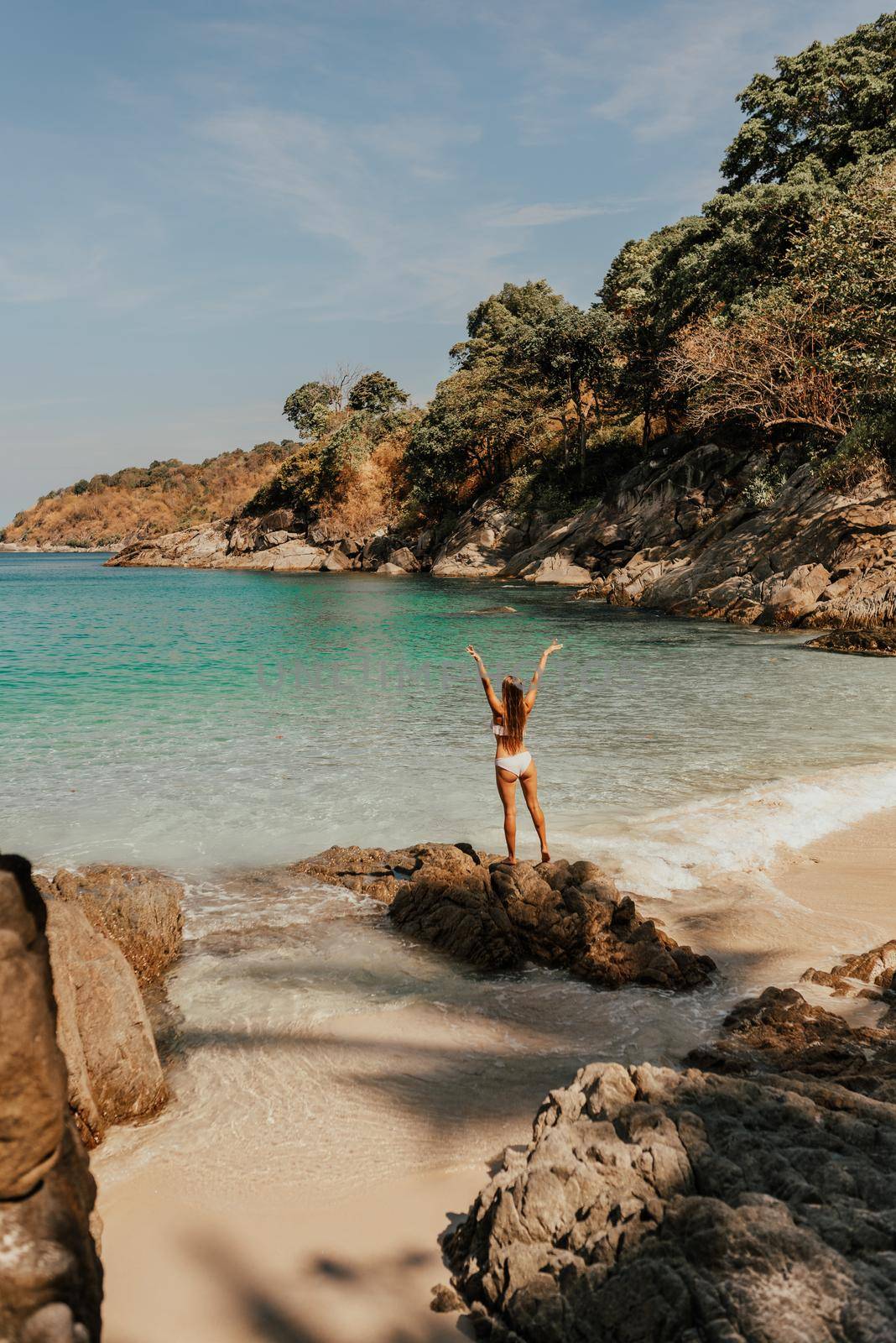 a young blonde European woman in a white bikini swimsuit on the beach stands backwards on the stone rocks on the ocean shore.