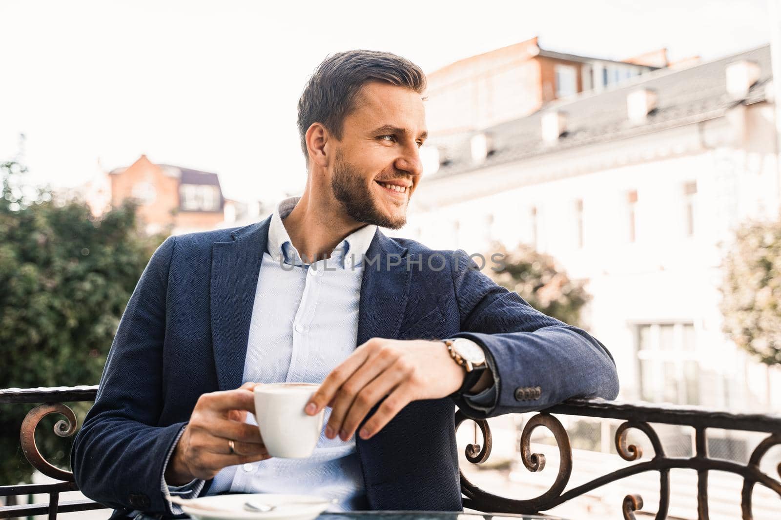 Man is sitting on the summer terrace in cafe, drinking coffee and smiling. Handsome man with cup of coffee in cafe. Morning lifestyle of male