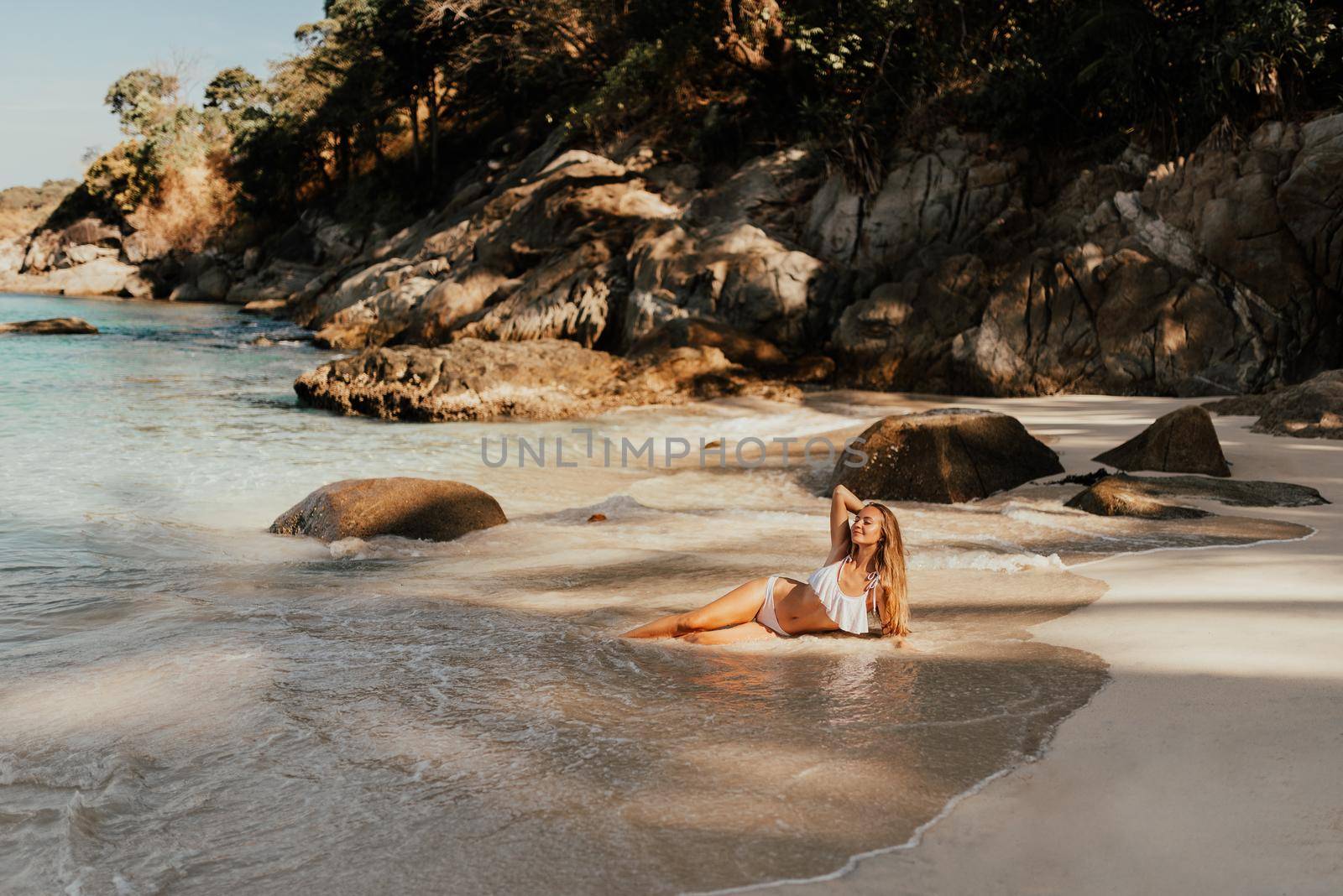 young blond European woman in white bikini swimsuit on beach lies on sand beach sea. background of large stone rocks and palm trees