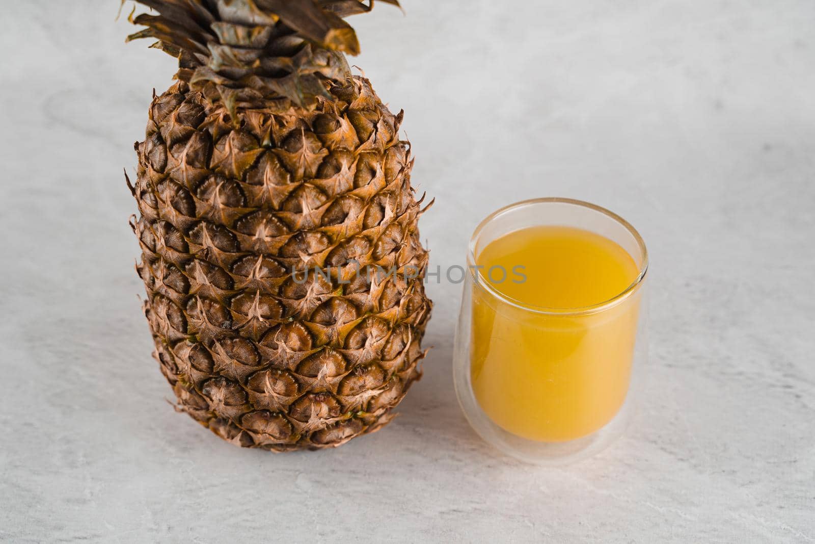 Pineapple fruit and juice in double glass cup on white stone background. Pouring yellow tropical fruit juice into glass. by Rabizo