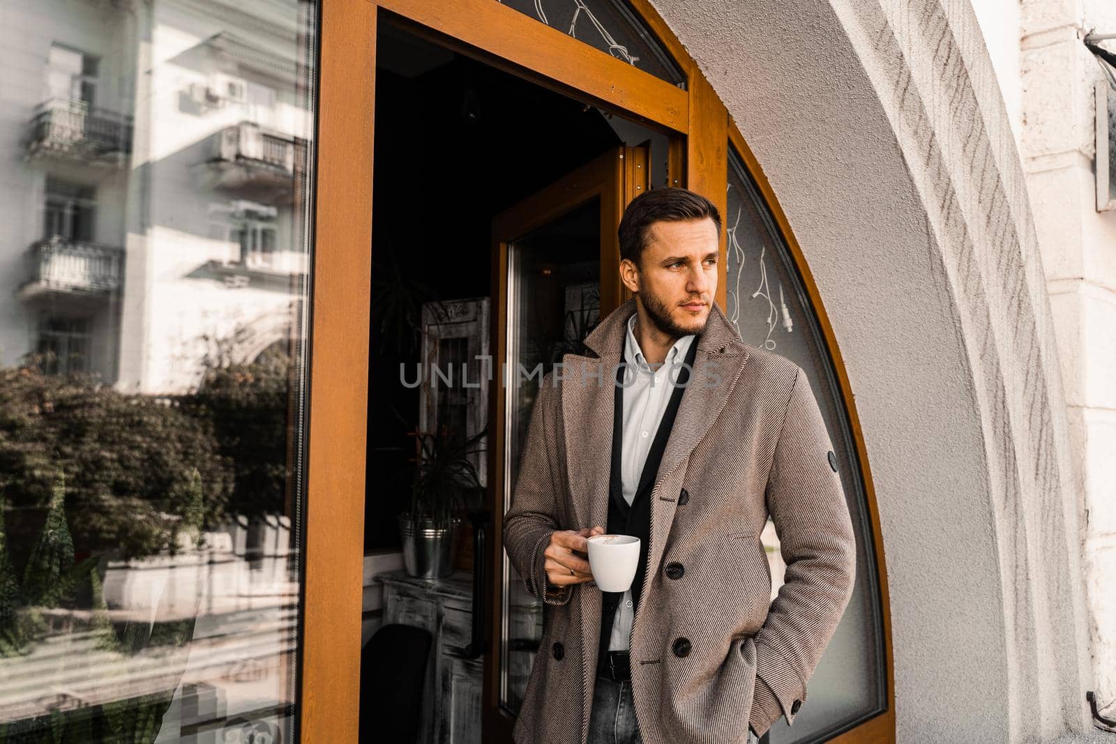 Handsome man with cup of coffee posing in coat in cafe. Male lifestyle. by Rabizo