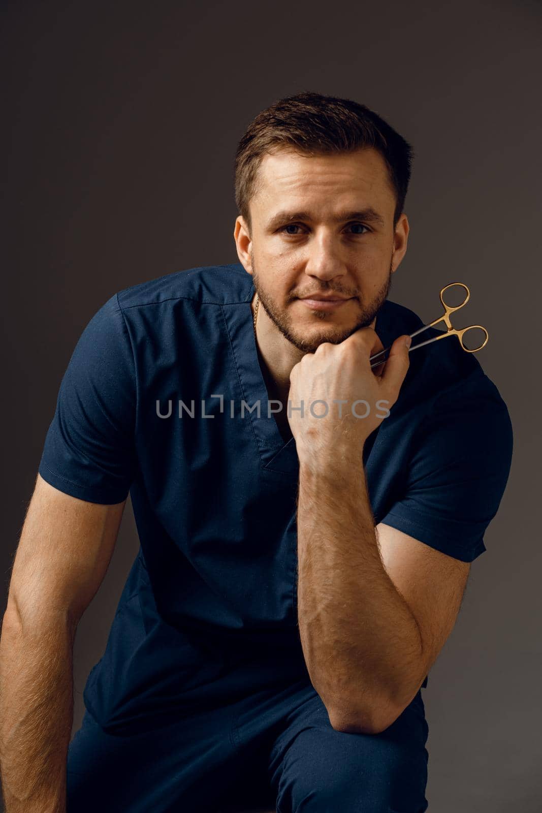 Handsome doctor with surgical scissors close-up. Confident man holding medical equipment in hands and smiling. Happy male posing in studio. by Rabizo