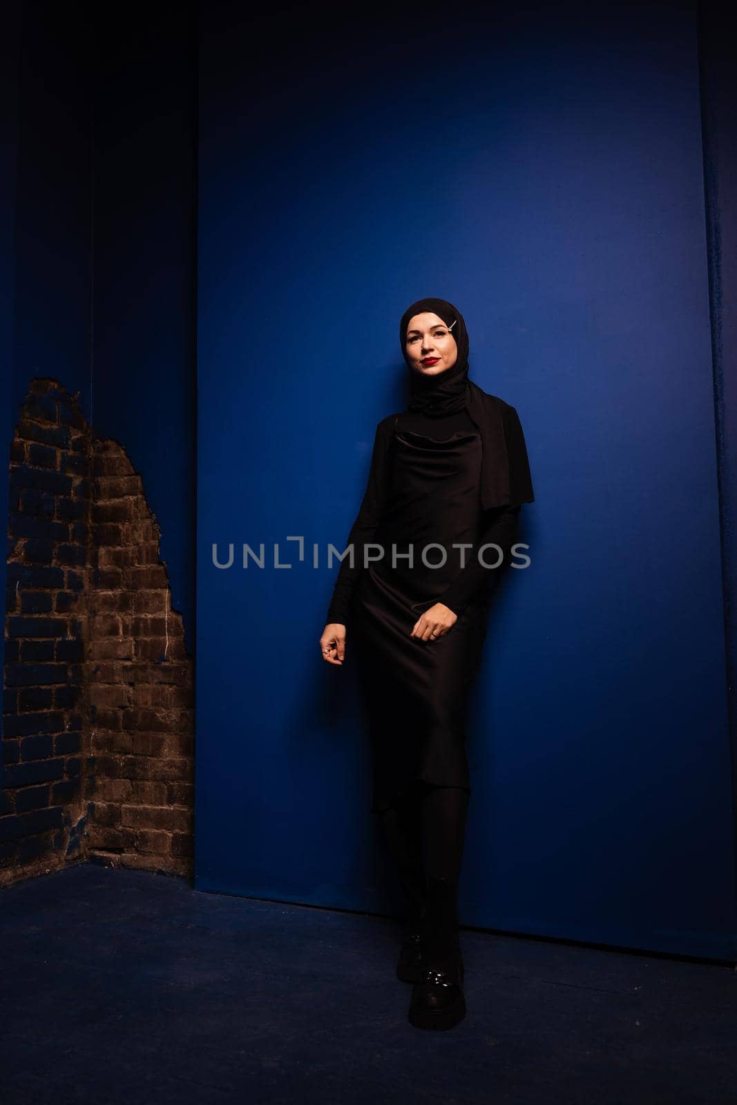 Fashion muslim woman in black hijab is posing on blue background in studio. Professional caucasian model