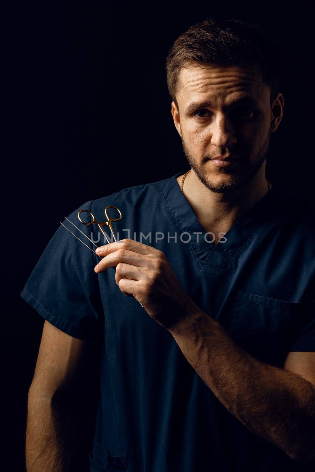 Handsome doctor with surgical scissors on dark background. Confident man holding medical equipment in hands and smiling. Happy male posing in studio