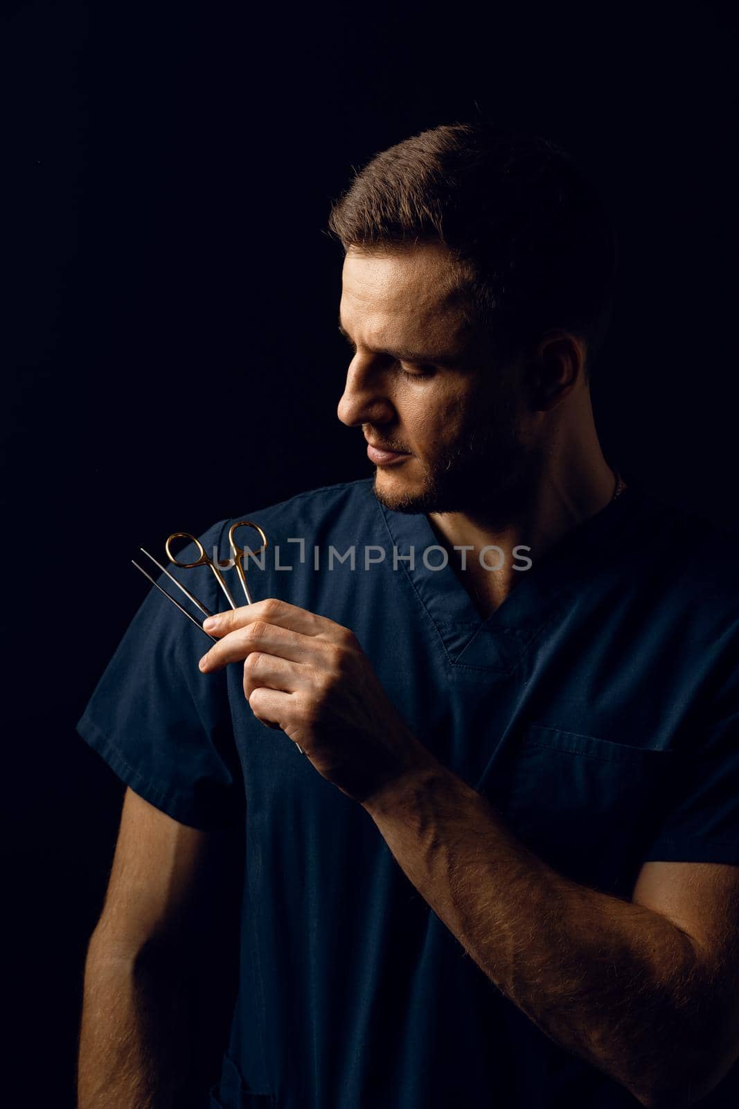 Handsome doctor with surgical scissors on dark background. Confident man holding medical equipment in hands and smiling. Happy male posing in studio. by Rabizo