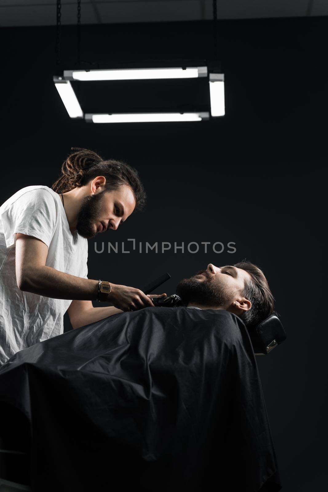 Beard shaving in barbershop. Barber with dreadlocks trim handsome bearded man. by Rabizo