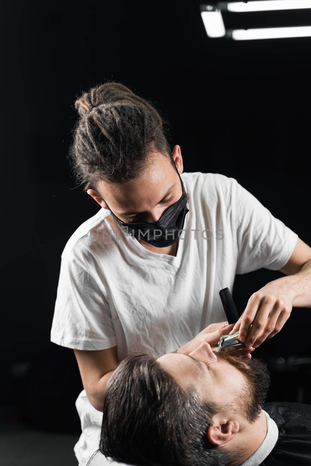 Mustache shaving in barbershop. Barber with dreadlocks in black medical mask trim beard of handsome man at quarantine coronavirus covid-19. by Rabizo