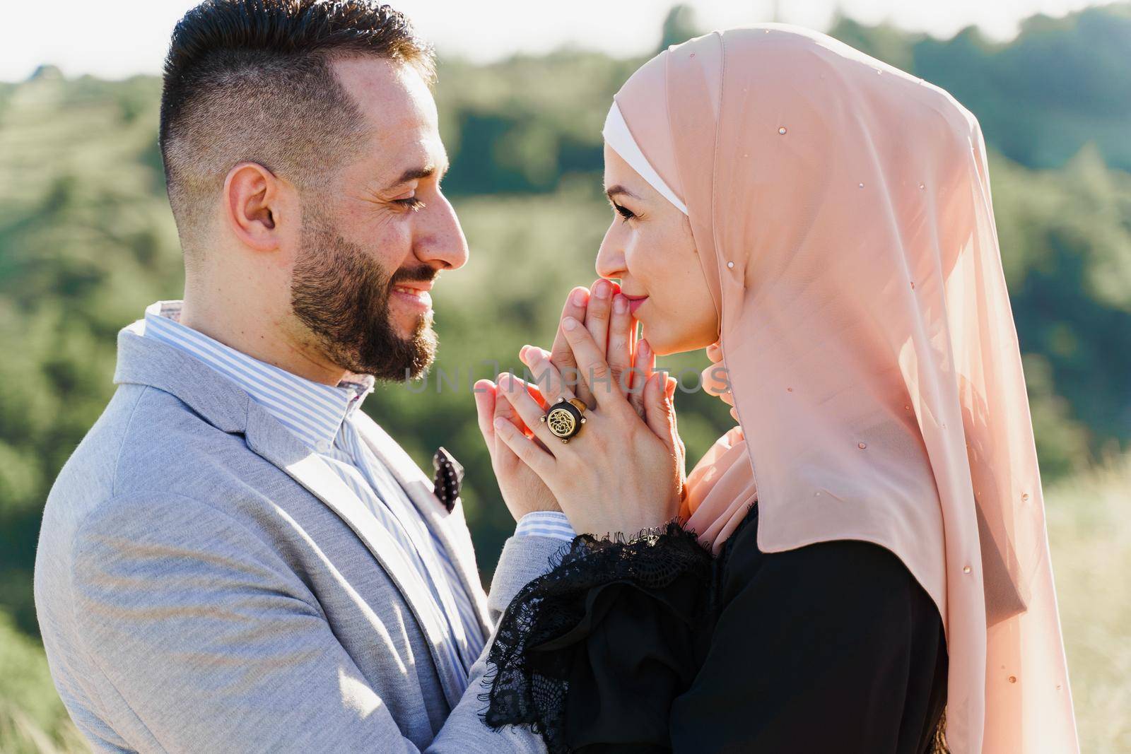 Muslim love story close-up. Mixed couple smiles and hugs on the green hills . Woman weared in hijab looks to her man. Advert for on-line dating agency