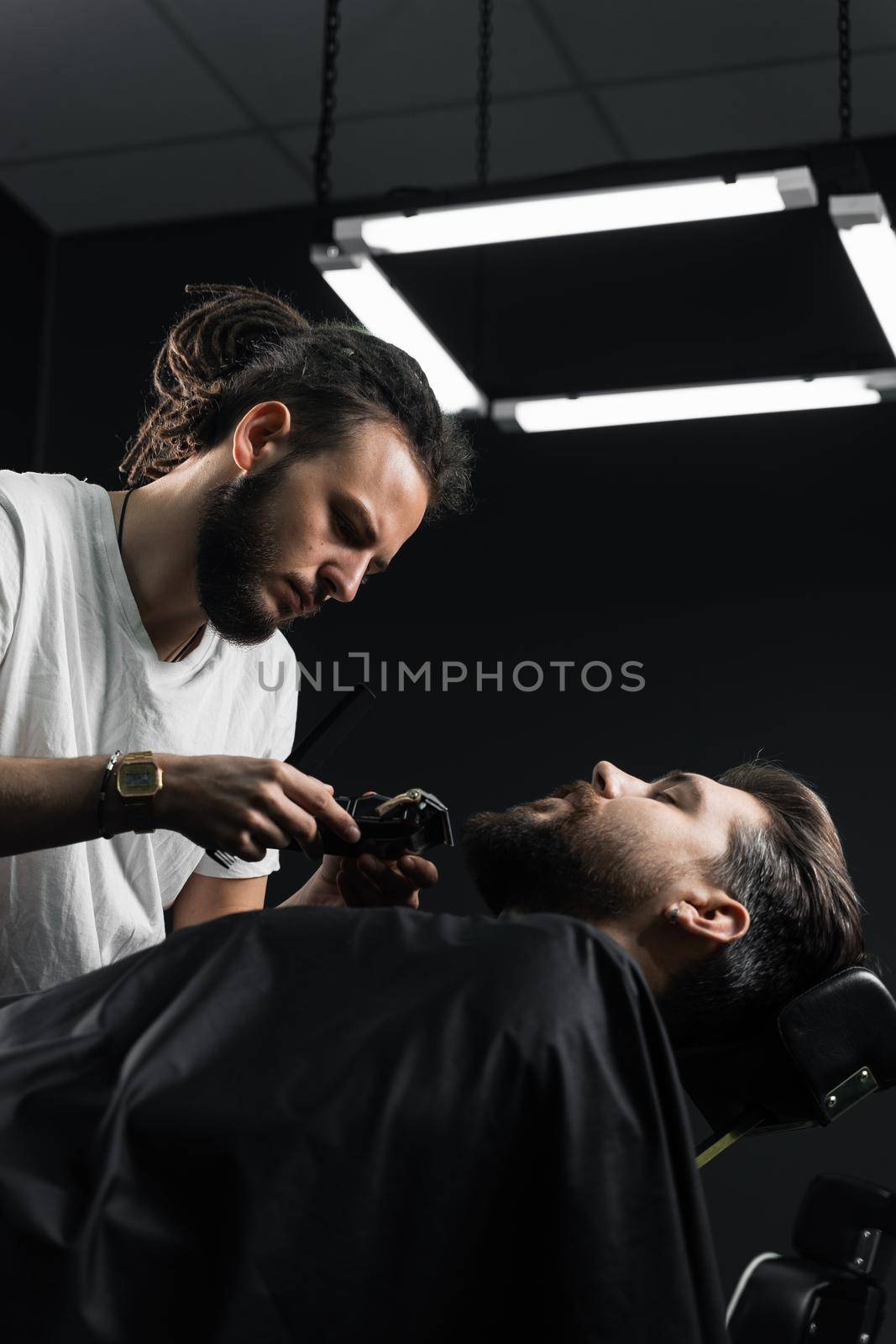 Handsome bearded man has hair and beard cut in barbershop. Dreadlock barber cuts client's hair.