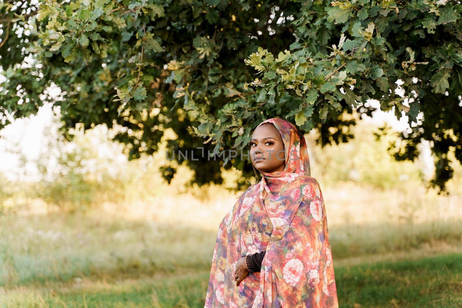 Muslim black woman African ethnicity weared traditional colorful hijab smiles and stay under green tree. Pretty black girl smiles.