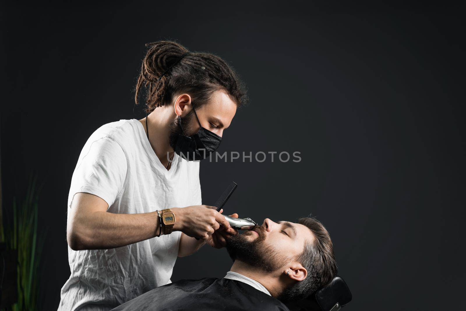 Mustache shaving in barbershop. Barber with dreadlocks in black medical mask trim beard of handsome man at quarantine coronavirus covid-19. by Rabizo