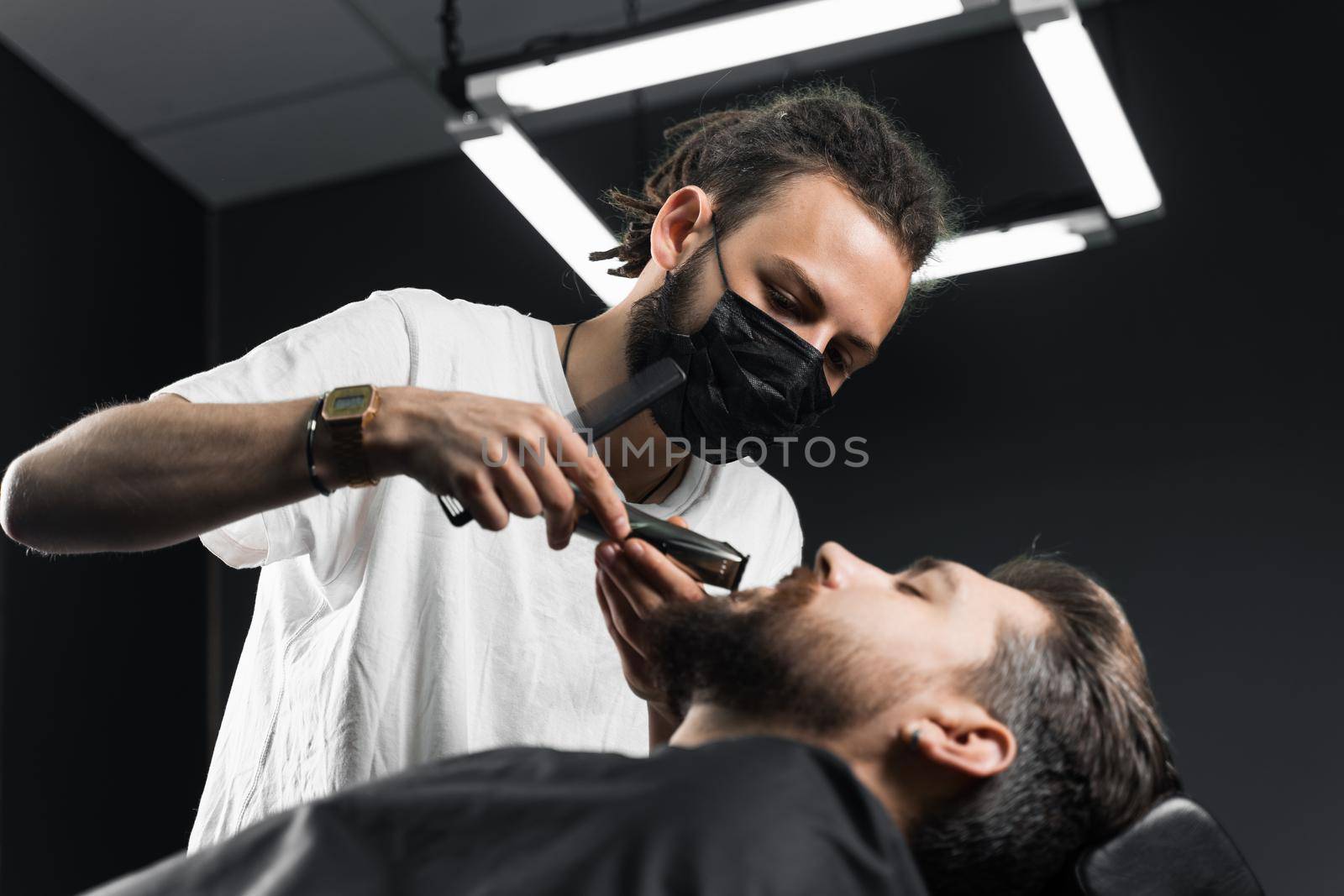 Mustache shaving in barbershop. Barber with dreadlocks in black medical mask trim beard of handsome man at quarantine coronavirus covid-19. by Rabizo