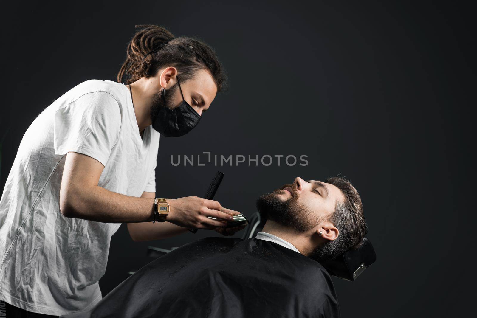 Beard shaving in barbershop. Barber with dreadlocks in black medical mask trim bearded man at quarantine coronavirus covid-19