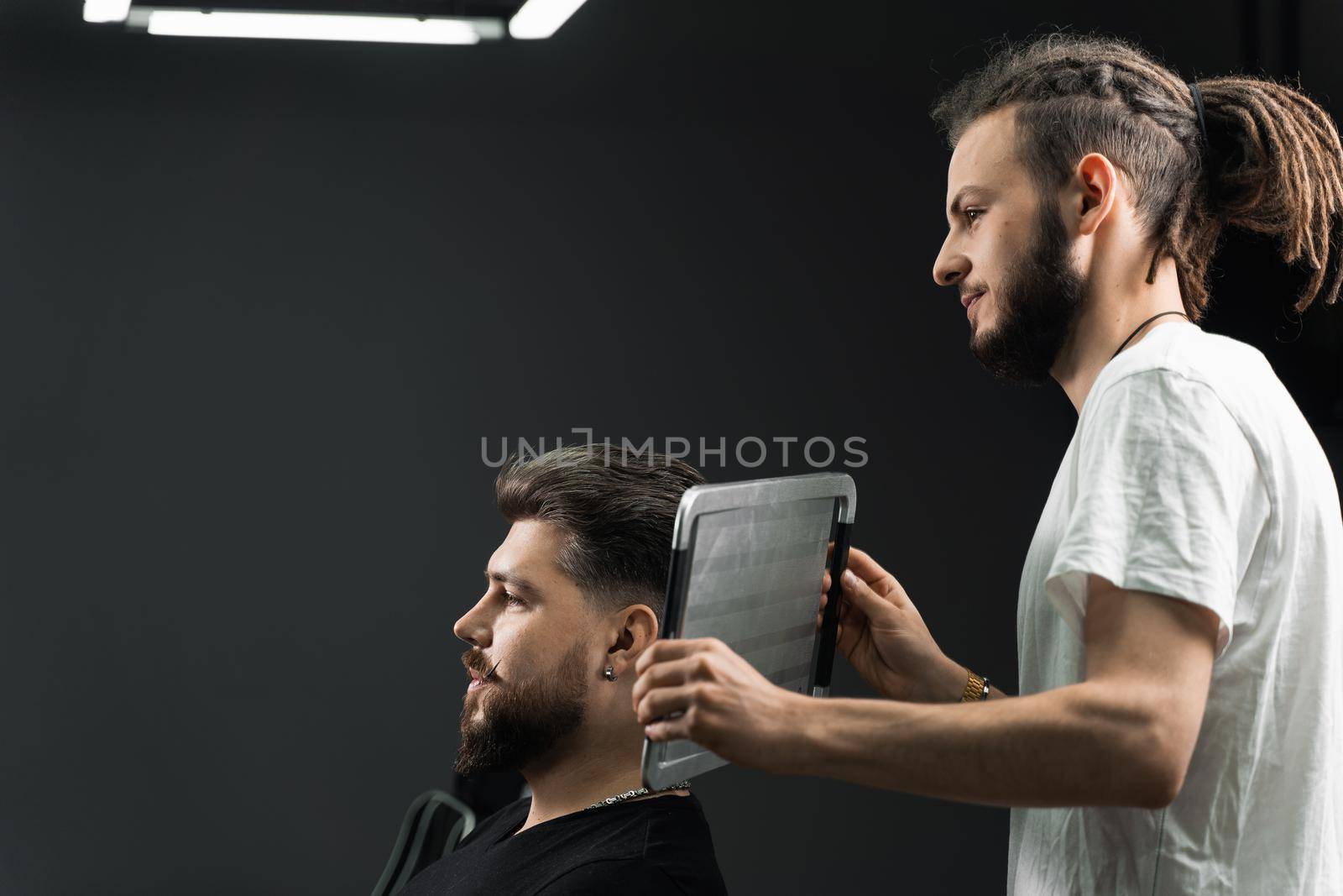 Barber with dreadlocks showing result of low fade machine hair and beard cut for bearded man in barbershop. Hairstyle with a smooth transition